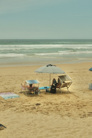 A sandy beach with two people lounging on deck chairs under umbrellas. The ocean waves gently roll in the background, creating a serene and relaxing atmosphere. Various beach towels are spread out nearby.
