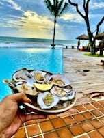 A hand holds a plate of oysters on ice, garnished with herbs and a lemon wedge, against the backdrop of a serene poolside setting. The pool borders a wooden deck that leads to a sandy beach with gentle ocean waves under a partly cloudy sky. Palm trees and thatched umbrellas add to the tropical ambiance.