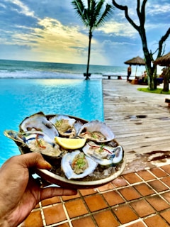 A hand holds a plate of oysters on ice, garnished with herbs and a lemon wedge, against the backdrop of a serene poolside setting. The pool borders a wooden deck that leads to a sandy beach with gentle ocean waves under a partly cloudy sky. Palm trees and thatched umbrellas add to the tropical ambiance.