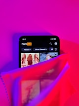 A smartphone screen displaying a website with an adult content theme partially covered by a sheer fabric in vibrant lighting. The colors are predominantly pink and purple, with some red hues. The website interface shows categories like 'Photos' and 'Most Viewed.'
