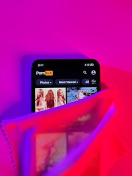 A smartphone screen displaying a website with an adult content theme partially covered by a sheer fabric in vibrant lighting. The colors are predominantly pink and purple, with some red hues. The website interface shows categories like 'Photos' and 'Most Viewed.'