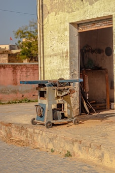 A vintage machinery piece, possibly a woodworking or metalworking tool, stands outside an old, weathered building. The machine is mounted on wheels and has a blue and silver finish. The building's faded yellow exterior and slightly ajar door reveal a dimly lit interior with tools hanging on the wall. The surroundings include a brick pathway and an older structure in the background.