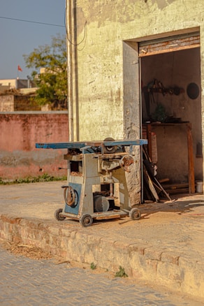 A vintage machinery piece, possibly a woodworking or metalworking tool, stands outside an old, weathered building. The machine is mounted on wheels and has a blue and silver finish. The building's faded yellow exterior and slightly ajar door reveal a dimly lit interior with tools hanging on the wall. The surroundings include a brick pathway and an older structure in the background.
