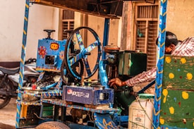 A vibrant street-side juice machine, decorated with colorful patterns and designs, is being operated by an unseen person in a red plaid shirt. Various components, including a wheel and levers, are visible, suggesting mechanical operation. A motor and a metal container with Pepsi branding are part of the setup. The backdrop includes blurred motorcycles and buildings, hinting at an urban environment.