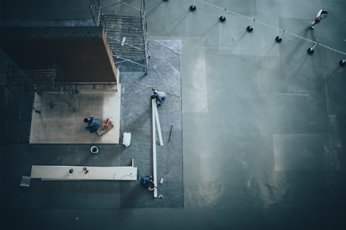A high-angle view of two workers, possibly in a construction or setup situation, with scaffolding and large wooden panels on a concrete surface. One person is kneeling while the other is standing, possibly installing or measuring materials. The space is open and industrial with visible tools and materials scattered, and a person walks in the distance behind a series of bollards connected with ropes.