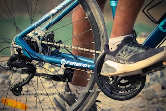 Close-up view of a bicycle's drivetrain and rear wheel with the brand name 'Firefox' visible on the frame. A person's leg and foot are on a pedal, wearing dark athletic shoes labeled 'Ultra-White.' The image is focused on the mechanical components, including the chain, gears, and derailleurs.