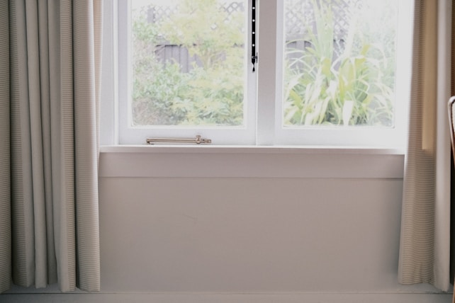 A bright room features a large window framed by light-colored curtains, looking out onto a garden with green foliage. The window is double-paned and set in a white frame. Sunlight filters through, creating a serene atmosphere.