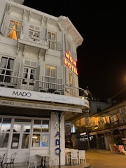 A corner building with a classic architectural style is illuminated at night. The building features a neon sign reading 'MOM HOTEL' and downstairs is a cafe called 'Mado Dondurma Cafe'. There are tables and chairs outside and the street is quiet.