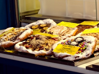 An assortment of pastries displayed on a counter, each topped with varied ingredients like sliced bananas, powdered sugar, nuts, chocolate shavings, and colorful candy-coated chocolates. They are accompanied by labels describing each type.