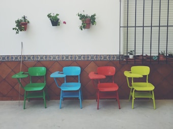 Four colorful chairs with attached writing desks are lined up against a wall. Each chair is a different color: green, blue, red, and yellow. Above them, potted plants are mounted on the wall. The wall is decorated with brown tiles featuring a blue pattern.