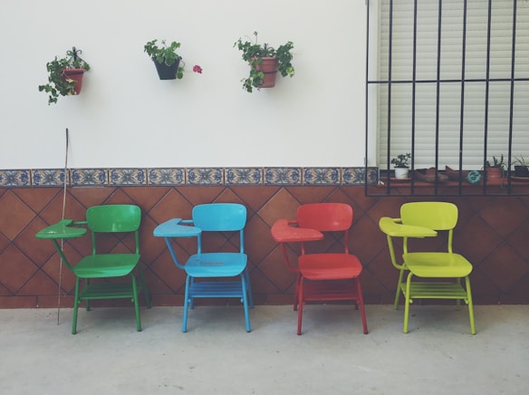 Four colorful chairs with attached writing desks are lined up against a wall. Each chair is a different color: green, blue, red, and yellow. Above them, potted plants are mounted on the wall. The wall is decorated with brown tiles featuring a blue pattern.