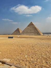 Two large pyramids stand prominently on a vast desert landscape under a clear blue sky with scattered clouds. Several vehicles, including a bus and a car, are visible on the sandy road in front of the pyramids.