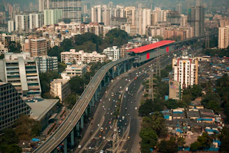 An urban landscape featuring a busy highway flanked by tall residential and commercial buildings. A metro rail with a red roof runs parallel to the highway on an elevated track. Dense clusters of high-rise buildings characterize the background, while greenery is visible in patches near the structures.