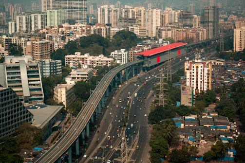 An urban landscape featuring a busy highway flanked by tall residential and commercial buildings. A metro rail with a red roof runs parallel to the highway on an elevated track. Dense clusters of high-rise buildings characterize the background, while greenery is visible in patches near the structures.