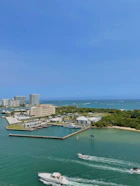 The image depicts a coastal cityscape with a marina and multiple buildings adjacent to a clear, blue body of water. Boats are navigating through the waterway, and patches of greenery can be seen on the shore.