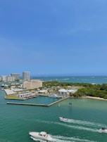 The image depicts a coastal cityscape with a marina and multiple buildings adjacent to a clear, blue body of water. Boats are navigating through the waterway, and patches of greenery can be seen on the shore.