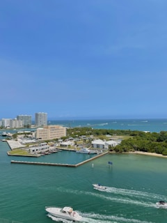 The image depicts a coastal cityscape with a marina and multiple buildings adjacent to a clear, blue body of water. Boats are navigating through the waterway, and patches of greenery can be seen on the shore.