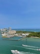 The image depicts a coastal cityscape with a marina and multiple buildings adjacent to a clear, blue body of water. Boats are navigating through the waterway, and patches of greenery can be seen on the shore.