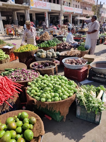 A bustling outdoor market scene with a variety of fresh vegetables arranged in baskets and crates. There are people interacting and browsing through the produce, which includes lemons, cucumbers, carrots, and squash, among others. The market stalls are set against a backdrop of street shops and buildings.