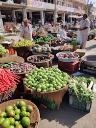 A bustling outdoor market scene with a variety of fresh vegetables arranged in baskets and crates. There are people interacting and browsing through the produce, which includes lemons, cucumbers, carrots, and squash, among others. The market stalls are set against a backdrop of street shops and buildings.