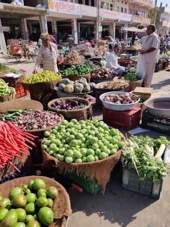 A bustling outdoor market scene with a variety of fresh vegetables arranged in baskets and crates. There are people interacting and browsing through the produce, which includes lemons, cucumbers, carrots, and squash, among others. The market stalls are set against a backdrop of street shops and buildings.