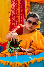 A woman wearing sunglasses and traditional attire is engaged in a ceremonial activity. She is pouring water from a colorful pot into a small pool, surrounded by vibrant marigold garlands. The background is decorated with bright, colorful fabrics and floral patterns.