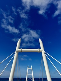 A modern architectural structure resembling a bridge or tower with sleek, white beams and cables set against a vivid blue sky with scattered clouds.