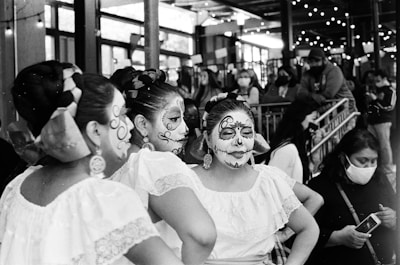 People with intricate face paint and traditional dresses gather in a festive setting decorated with string lights. They appear to be part of a cultural or ceremonial event, surrounded by other onlookers, some wearing masks.