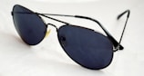 A pair of dark-lensed aviator sunglasses with thin metal frames, resting on a white surface. The lenses are tinted and reflective, with a classic design featuring a double bridge.
