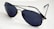A pair of dark-lensed aviator sunglasses with thin metal frames, resting on a white surface. The lenses are tinted and reflective, with a classic design featuring a double bridge.