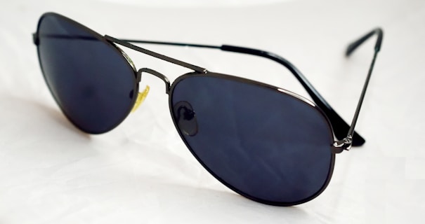 A pair of dark-lensed aviator sunglasses with thin metal frames, resting on a white surface. The lenses are tinted and reflective, with a classic design featuring a double bridge.