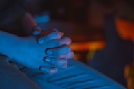 A close-up view of two hands clasped together, illuminated by a blue light. The soft focus and subdued lighting create a sense of calm and introspection.