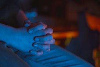 A close-up view of two hands clasped together, illuminated by a blue light. The soft focus and subdued lighting create a sense of calm and introspection.