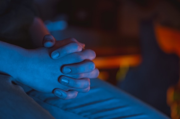 A close-up view of two hands clasped together, illuminated by a blue light. The soft focus and subdued lighting create a sense of calm and introspection.