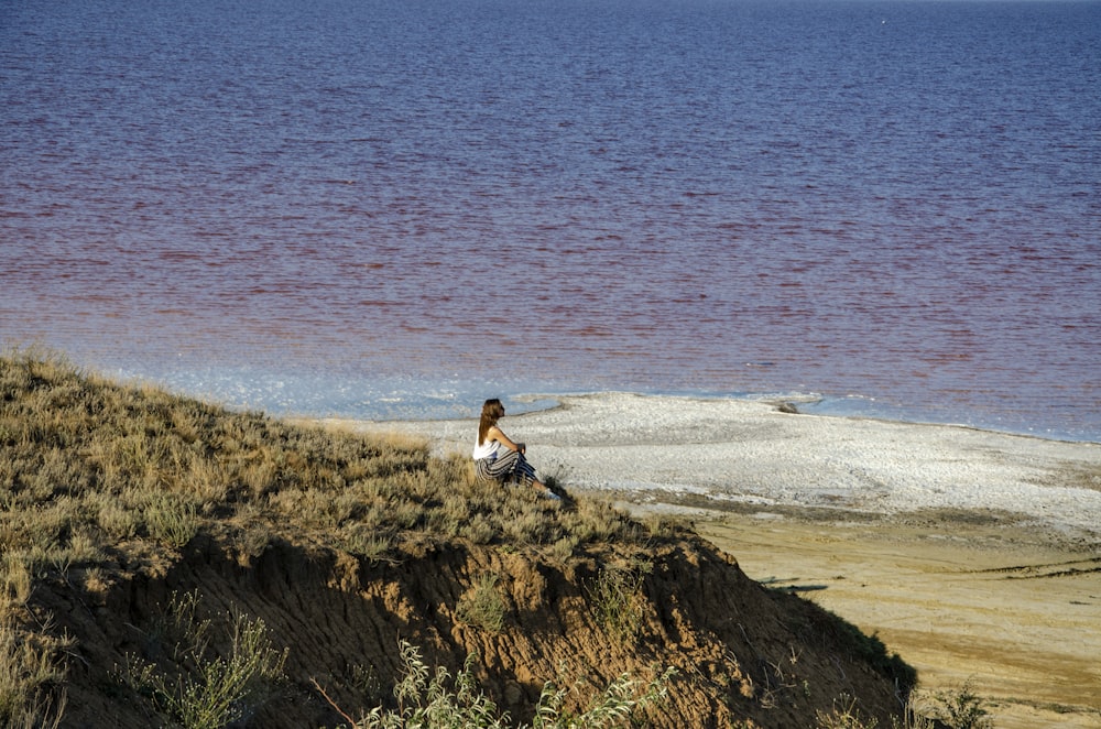 a woman sitting on top of a hill next to a body of water