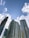 Tall, modern skyscrapers tower against a bright blue sky dotted with white clouds. One building features a unique curved design and another with a grid-like facade. A construction crane is visible, indicating ongoing development.