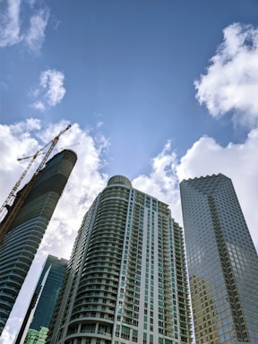 Tall, modern skyscrapers tower against a bright blue sky dotted with white clouds. One building features a unique curved design and another with a grid-like facade. A construction crane is visible, indicating ongoing development.