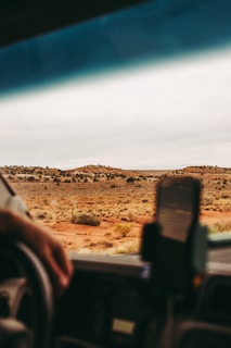 A perspective from inside a vehicle looking out at a vast desert landscape with sparse vegetation and distant low hills. The foreground shows part of the car's dashboard, a person's hand on the steering wheel, and a smartphone mounted on the dashboard.