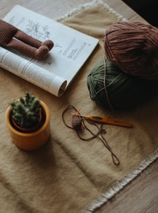 A cozy crafting setup featuring a rolled magazine, a crocheted doll, some yarn balls in brown and green, a crochet hook, and a small potted cactus on a table with a textured cloth.