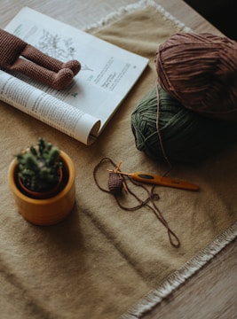 A cozy crafting setup featuring a rolled magazine, a crocheted doll, some yarn balls in brown and green, a crochet hook, and a small potted cactus on a table with a textured cloth.