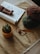 A cozy crafting setup featuring a rolled magazine, a crocheted doll, some yarn balls in brown and green, a crochet hook, and a small potted cactus on a table with a textured cloth.