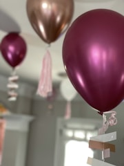 Several metallic balloons in shades of pink and gold are floating indoors with ribbons attached at the bottom. Soft lighting highlights their shiny surfaces and the surrounding neutral-colored walls offer a subtle backdrop.