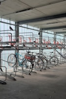 A bike rack inside a multi-level parking garage is shown with several bicycles parked. The structure is metallic with red accents on the upper tier, and pigeons are perched on the rack. The lighting is soft, and the area appears clean and organized.