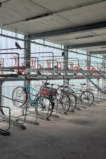 A bike rack inside a multi-level parking garage is shown with several bicycles parked. The structure is metallic with red accents on the upper tier, and pigeons are perched on the rack. The lighting is soft, and the area appears clean and organized.