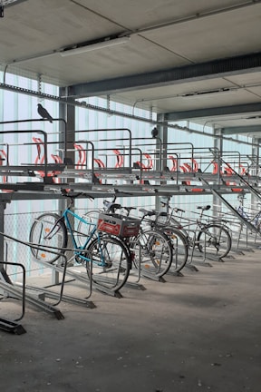 A bike rack inside a multi-level parking garage is shown with several bicycles parked. The structure is metallic with red accents on the upper tier, and pigeons are perched on the rack. The lighting is soft, and the area appears clean and organized.