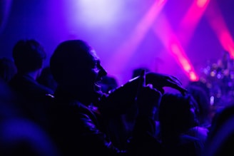 A lively concert scene with a crowd of people enjoying a live performance. Vivid shades of purple and pink stage lights illuminate the area, creating a dynamic and energetic atmosphere. A prominent silhouette of a person is visible in the foreground, possibly dancing or cheering.