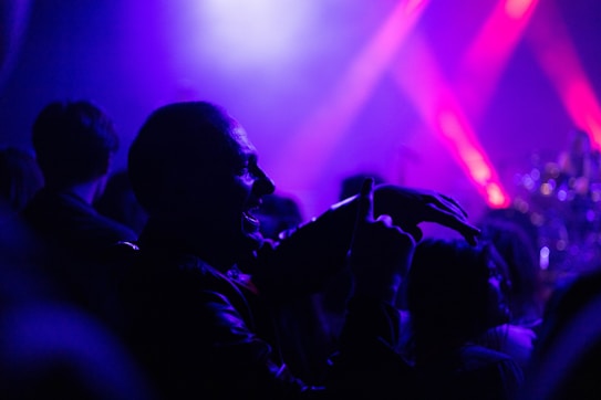 A lively concert scene with a crowd of people enjoying a live performance. Vivid shades of purple and pink stage lights illuminate the area, creating a dynamic and energetic atmosphere. A prominent silhouette of a person is visible in the foreground, possibly dancing or cheering.