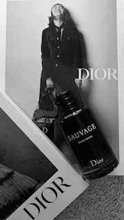 A black and white image featuring a Dior advertisement with a stylishly dressed person in a denim outfit, holding a handbag. In front of this photo is a bottle labeled 'Sauvage Eau de Parfum' by Dior. An open book with another word 'Dior' on the edge adds to the elegant aesthetic.