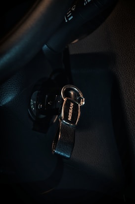 A key with a black leather strap and a metal clasp, featuring the brand name 'Nissan'. It is inserted into the ignition of a car, which is part of a dark interior, including sections of the steering wheel and dashboard.