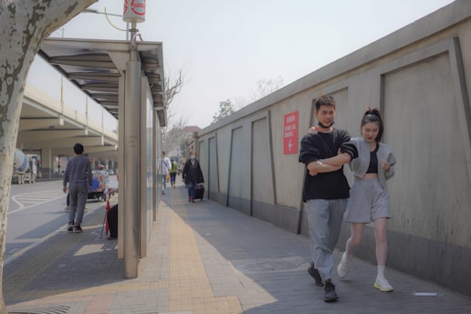 A young man and woman are walking together along a city sidewalk. The man has his arm around the woman, and they both appear relaxed and casual, dressed in comfortable clothes. Other pedestrians can be seen in the background, some carrying bags. The setting includes a road with vehicles, a bus stop, and a large concrete wall with a red sign.